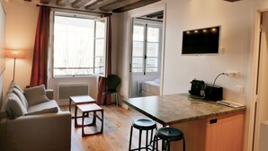 Un espacio habitable compacto con sofá, una pequeña mesa de comedor, una cocina con televisor y una ventana con cortinas.