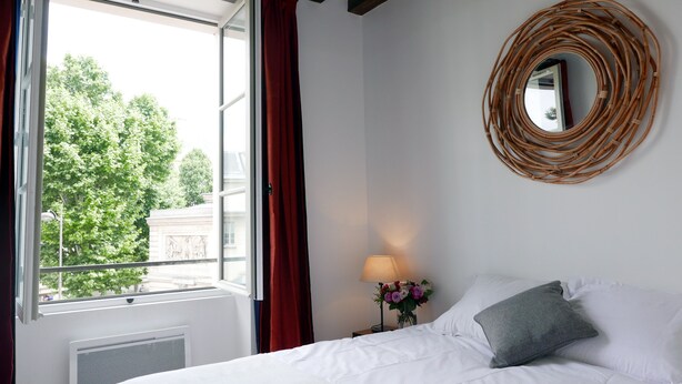 Habitación de hotel con cama, ventana con cortinas rojas, espejo en la pared, lámpara de noche y un jarrón con flores.