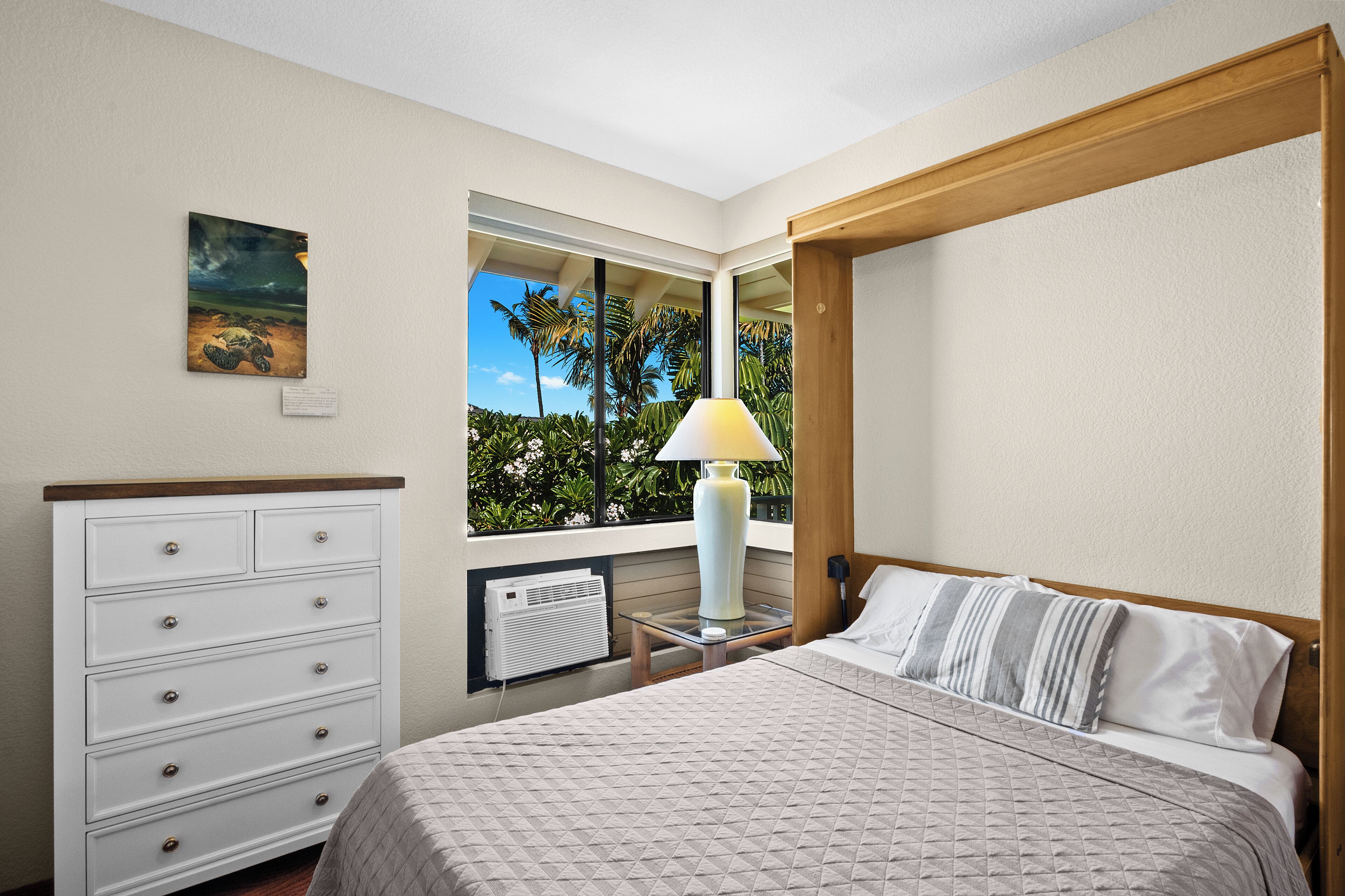 Une chambre à coucher avec un lit, une commode, une fenêtre donnant sur des palmiers et un tableau accroché au mur.