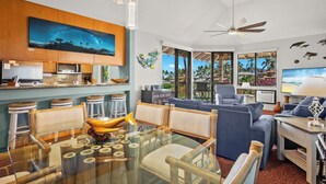 A modern living room with a glass dining table, blue sofas, and a large TV. - Sale!! Gorgeous 3BR 2BA Villa in Wailea, With Ocean, Golf and Sunset Views (Kihei)