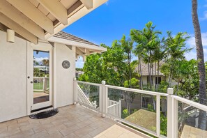 Uma varanda com grade branca, uma porta com painel de vidro e vista para uma área residencial com palmeiras e casas.
