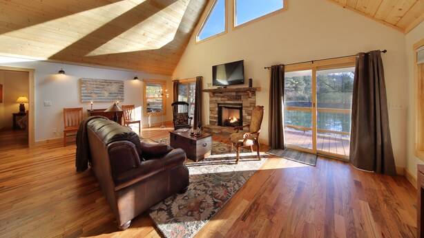 Un salón espacioso con suelos de madera, chimenea, sofá de cuero y grandes ventanales con vistas a un área exterior pintoresca.