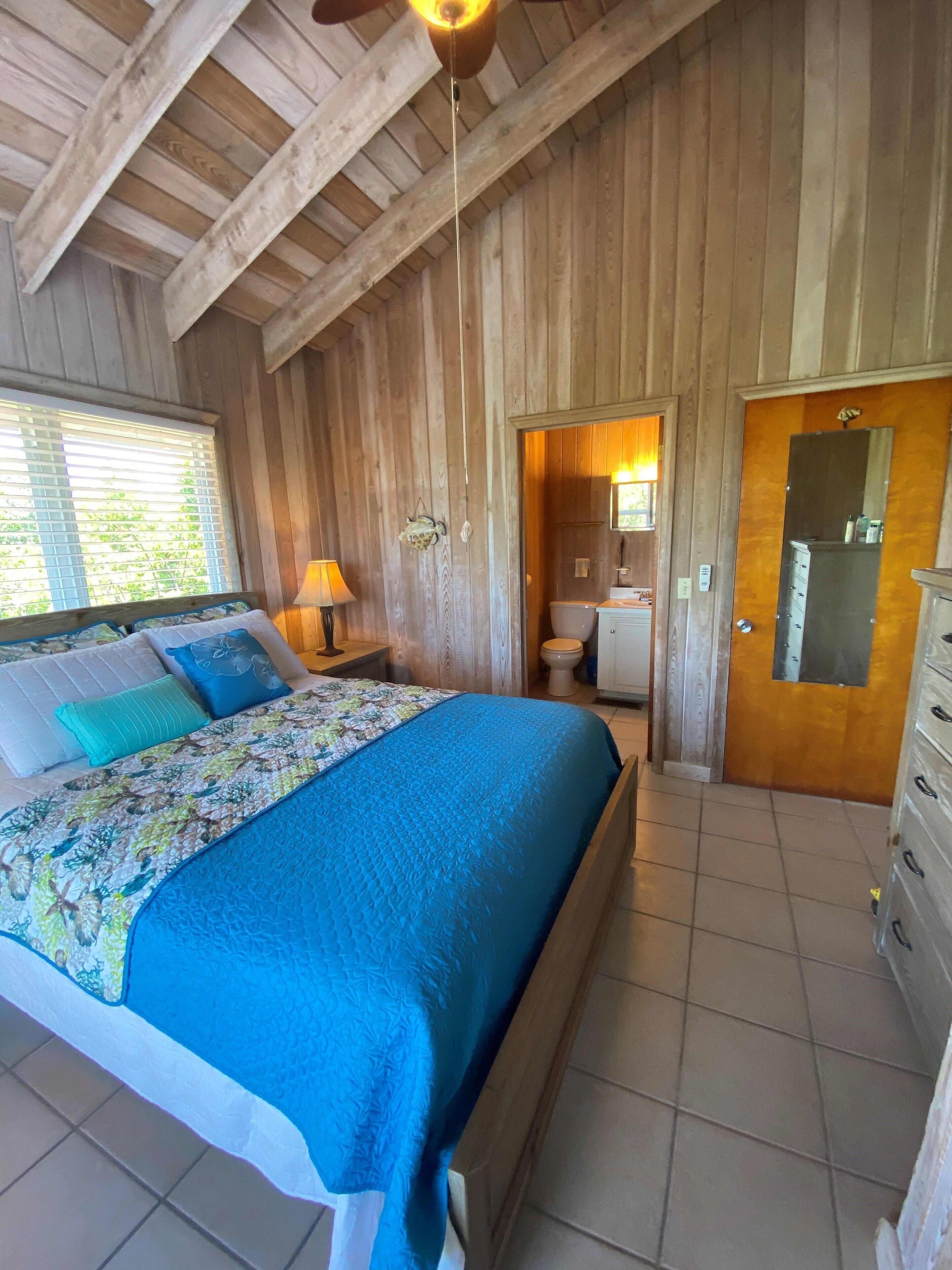 Une pièce en bois comprenant un lit, une table de chevet, une lampe, une porte et une salle de bain visible à travers une porte ouverte.