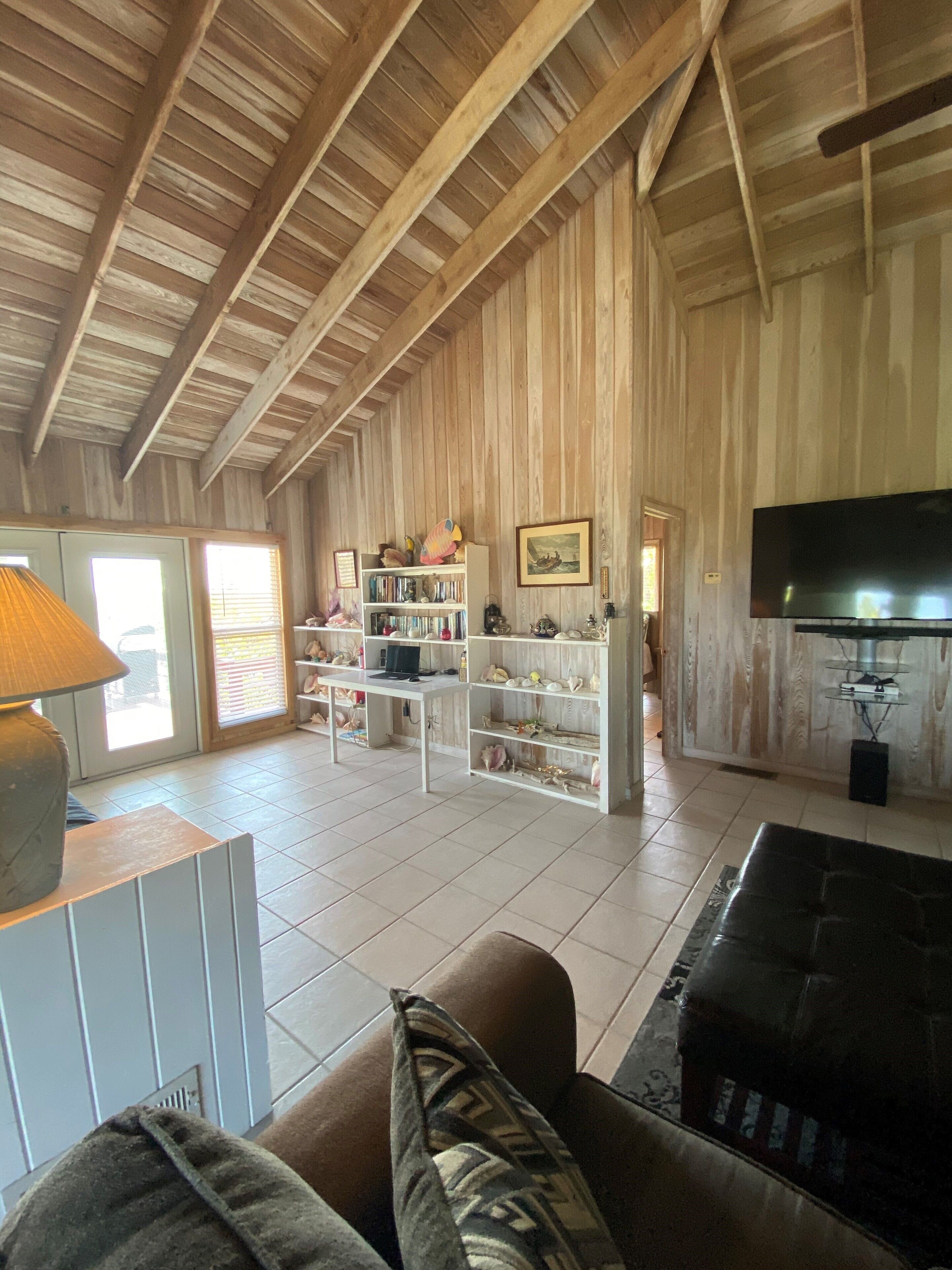 A spacious living area with a high ceiling, wooden beams, a large TV, and a bookshelf filled with decorative items.
