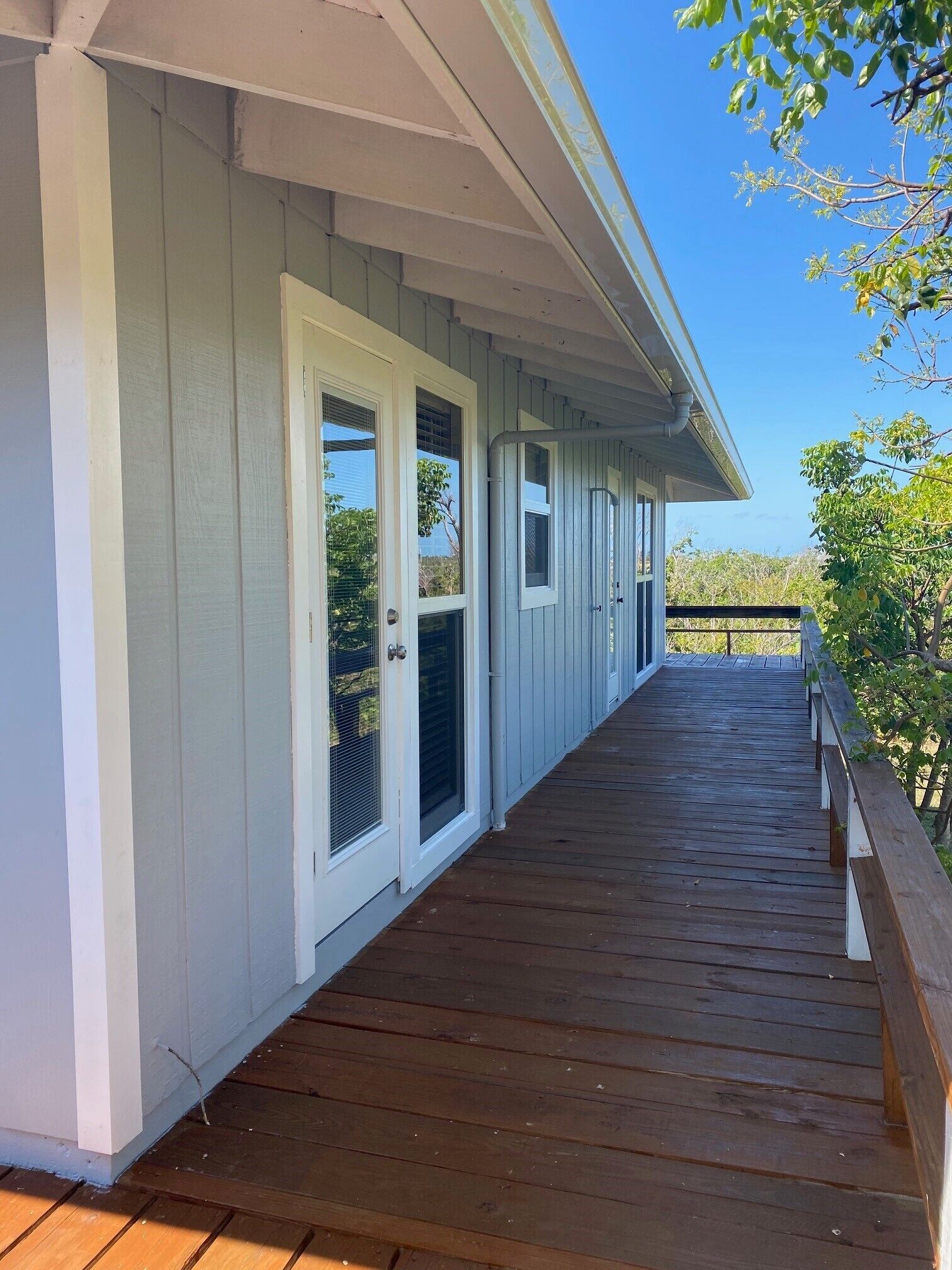 Une terrasse en bois couverte, avec un revêtement extérieur blanc et une balustrade, qui mène à un bâtiment doté de plusieurs portes et fenêtres.