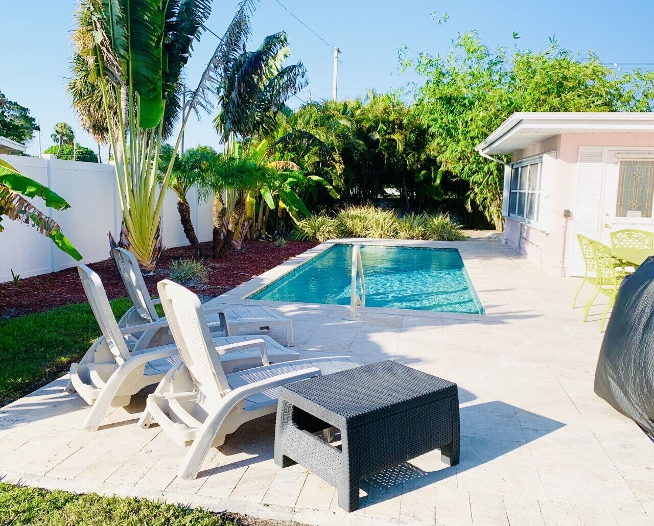 A pool area with lounge chairs and a small table, surrounded by tropical plants and a white fence.