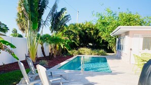 Uma área de piscina com espreguiçadeiras e uma pequena mesa, cercada por plantas tropicais e um muro branco.