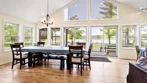 Uma sala de jantar com uma mesa grande, cadeiras, um candelabro e grandes janelas que oferecem vista para o exterior.