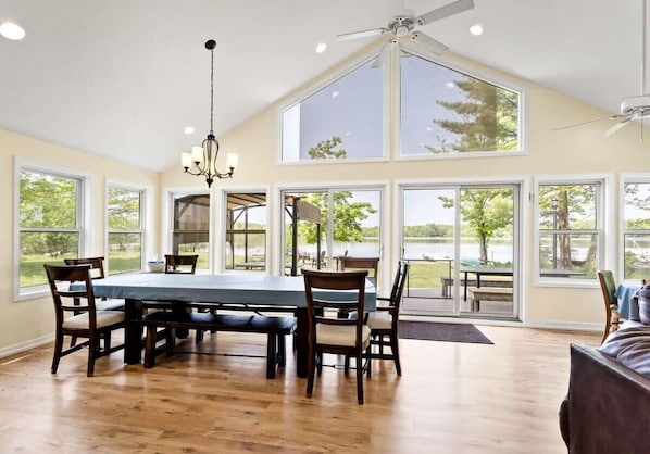 A dining area with a large table, chairs, a chandelier, and large windows offering views of the outdoors.