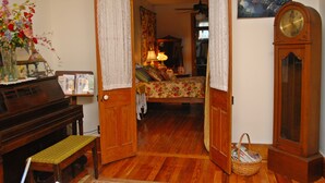 Una habitación con piso de madera, una cama con ropa de cama floral, un piano de madera, un reloj de pie y un cuadro en la pared.