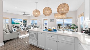 Un vaste espace de vie doté d’un grand îlot central, de meubles blancs, de luminaires suspendus en osier et d’un téléviseur diffusant l’émission « Cape & Coast ».