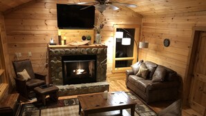 A cozy log cabin interior with a stone fireplace, leather sofas, a television, and a ceiling fan. - Buck Mountain Getaway - Stunning views, hot tub, fireplace. Nearby Boone. (Purlear)