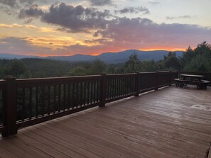 A wooden deck with a railing, overlooking a forested landscape and mountains during sunset. - Buck Mountain Getaway - Stunning views, hot tub, fireplace. Nearby Boone. (Purlear)