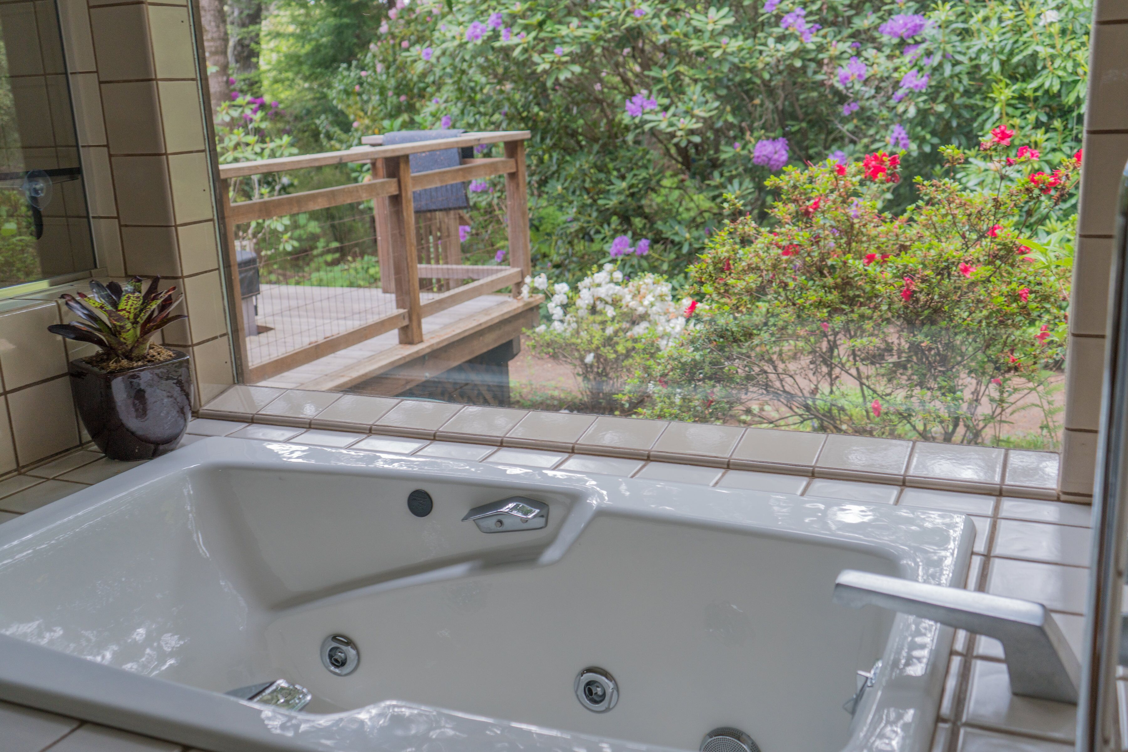 A bathroom with a bathtub, a potted plant, and a view of a garden with flowers and a wooden deck.