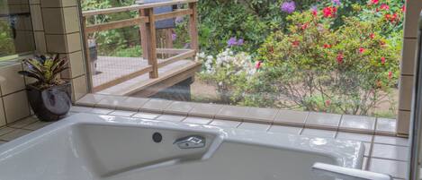 A bathroom with a bathtub, a potted plant, and a view of a garden with flowers and a wooden deck.