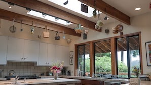 A modern kitchen with a large island, wooden beams, hanging baskets, and large windows offering a view of the outdoors. - Family Friendly Home on 20 Acre Farm (Caspar)