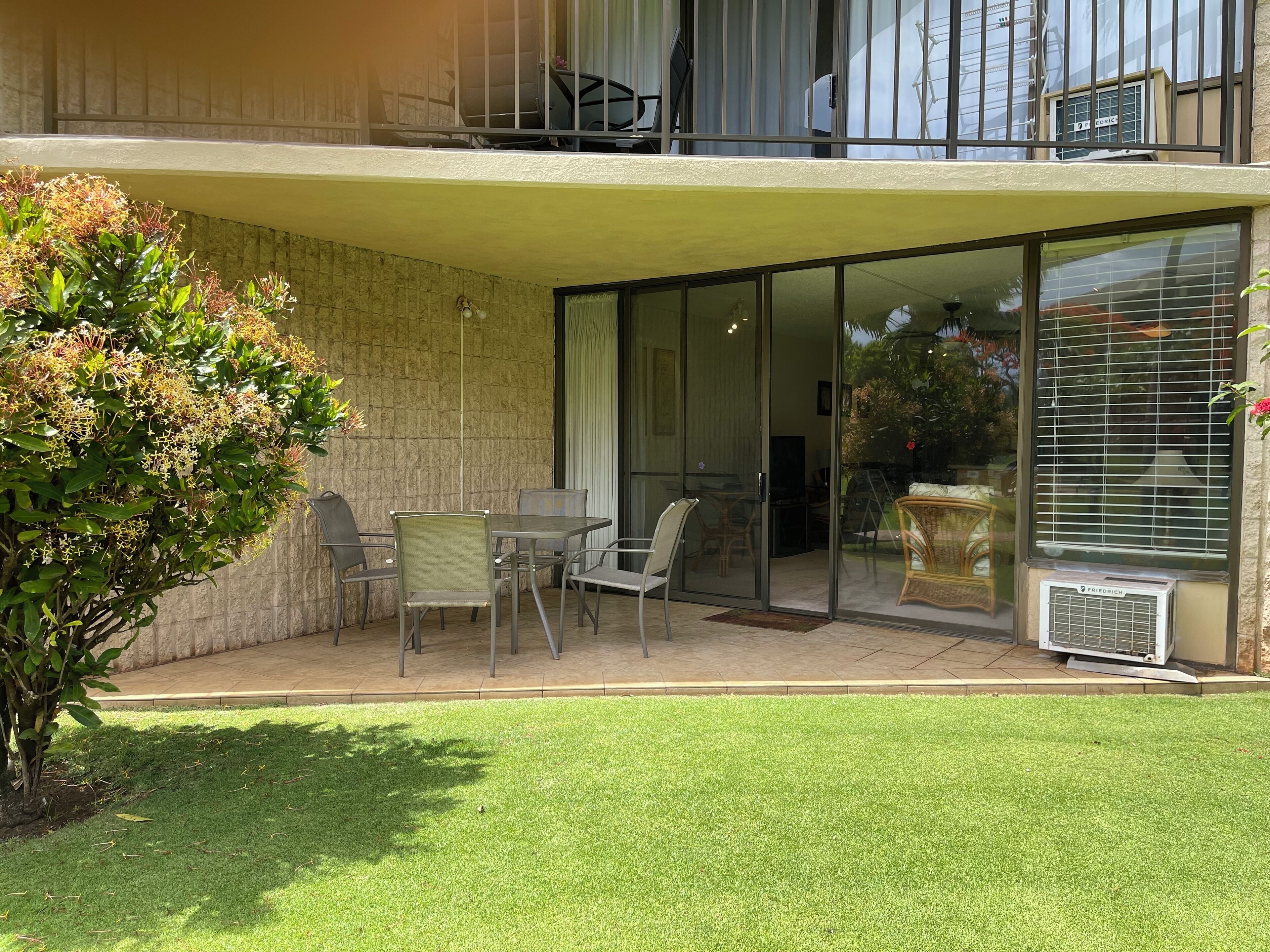 A two-story building with a covered patio area featuring a table and chairs, a small garden, and a sliding glass door leading to an interior space.