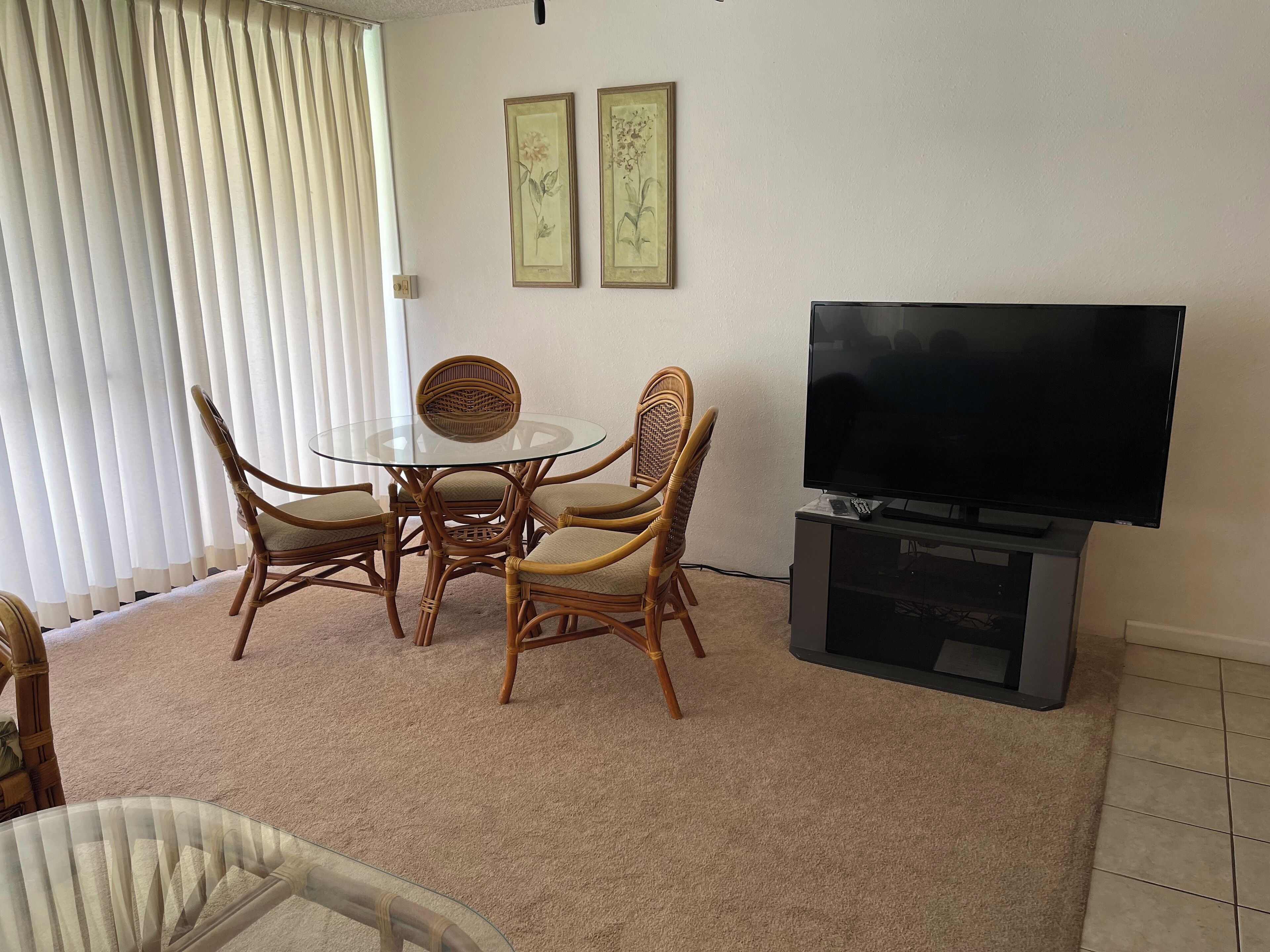 A living room with a round glass table surrounded by wicker chairs, a flat-screen TV on a stand, and two framed botanical prints on the wall.