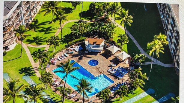 Luchtbeeld van een resort met een centraal gelegen zwembad, loungegedeeltes, palmbomen en een gebouw met een rood dak.
