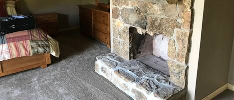 Un dormitorio con chimenea de piedra, una cama con manta a rayas, un tocador y una maleta en el suelo.