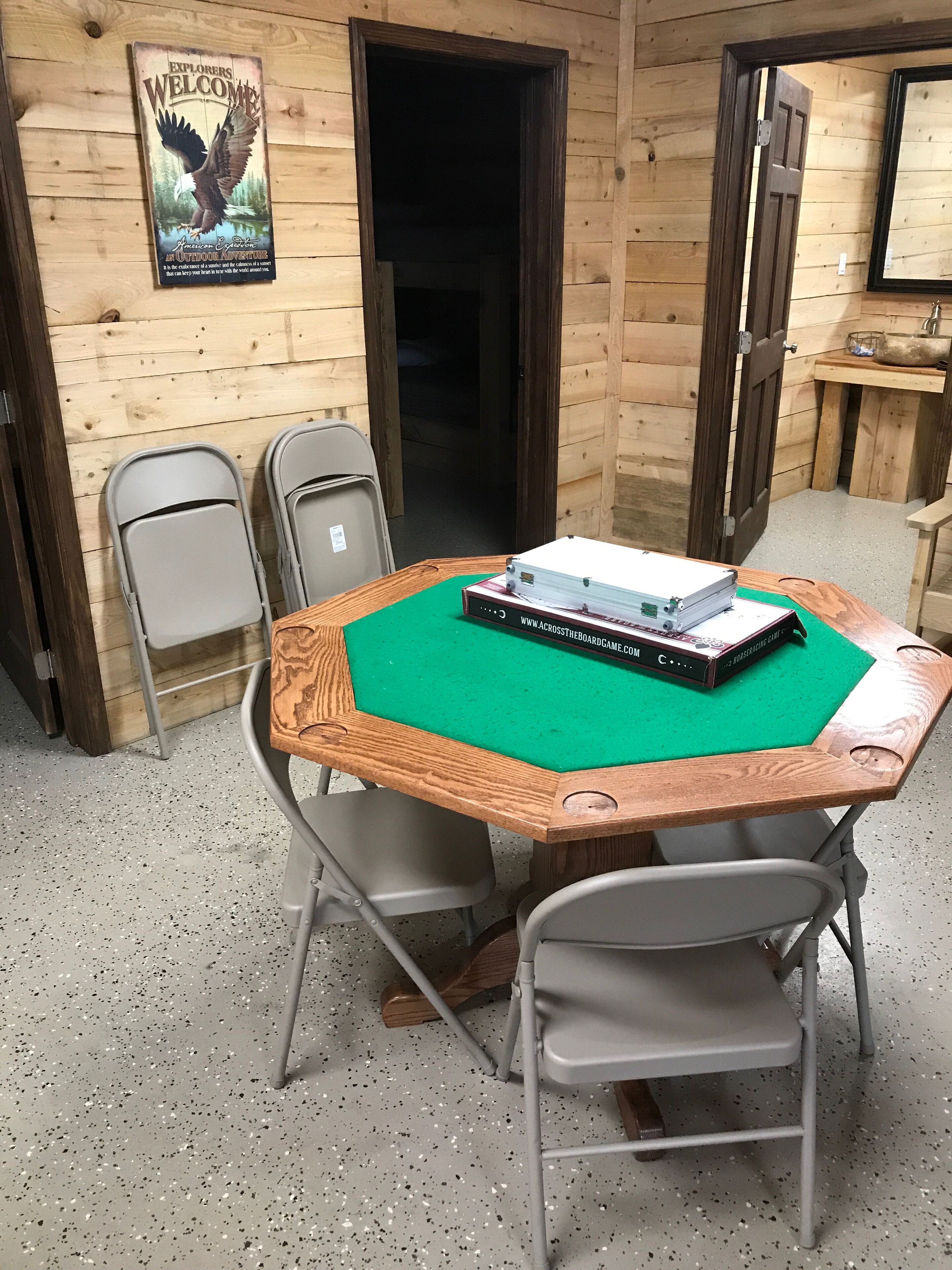 A wooden cabin interior with a green felt-covered table, two folding chairs, a wooden door, and a welcome sign.