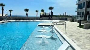 A swimming pool with clear blue water, surrounded by lounge chairs and palm trees, with a building in the background. - Beachside II 4338 - modern beachfront condo with pool, resort tram + balcony (Miramar Beach)