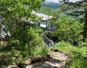 A stone staircase leading to a wooden deck with a metal railing, surrounded by lush greenery and overlooking a mountainous landscape. - Beautiful Mountain Home with Gorgeous View (Franklin)