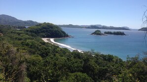 Un paysage côtier avec une plage, une végétation luxuriante et des collines à l’horizon.