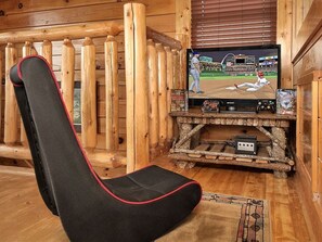 L’intérieur d’un chalet en bois est doté d’un téléviseur fixé sur un meuble rustique en bois, d’un fauteuil de jeu noir agrémenté de touches rouges et d’un plancher en bois.