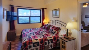 Un dormitorio con cama, suelo de madera, ventilador de techo, ventana y mesita de noche con lámpara.