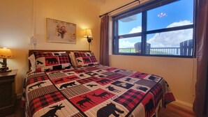 Un dormitorio con una cama grande, una ventana con vistas al cielo y nubes, una lámpara de pared y un cuadro en la pared.