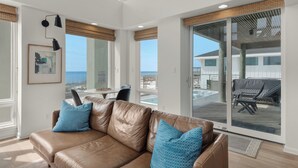A spacious living room with a brown leather sofa, a round table, and a large sliding glass door leading to a balcony with outdoor furniture.