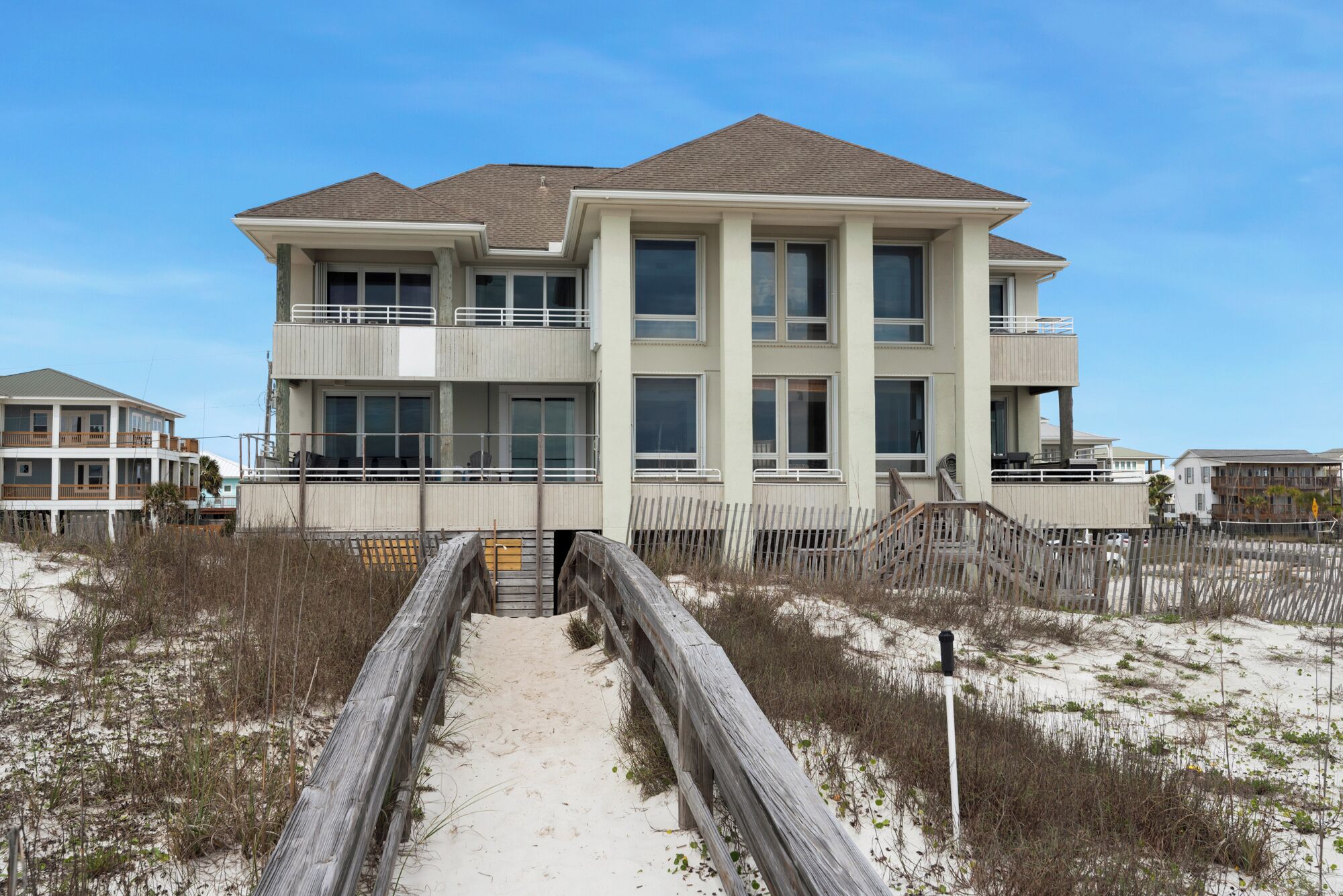 A two-story beachfront house with a sandy pathway leading to the entrance.