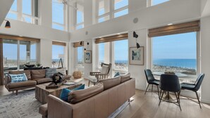 A spacious living room with high ceilings, large windows, and a view of the beach.