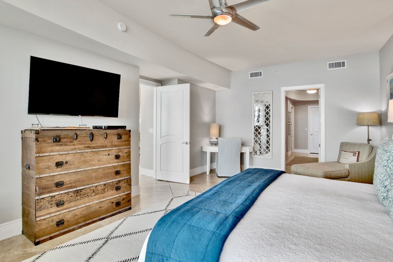 A bedroom with a bed, a wooden dresser, a television, a ceiling fan, and a door leading to another room.