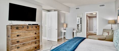 A bedroom with a bed, a wooden dresser, a television, a ceiling fan, and a door leading to another room.