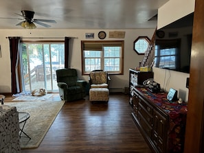 Relax in this inviting living room with a high ceiling fan, large windows offering a view of the outdoors, a green armchair, a floral-patterned chair, and a comfortable spot for your furry friend.