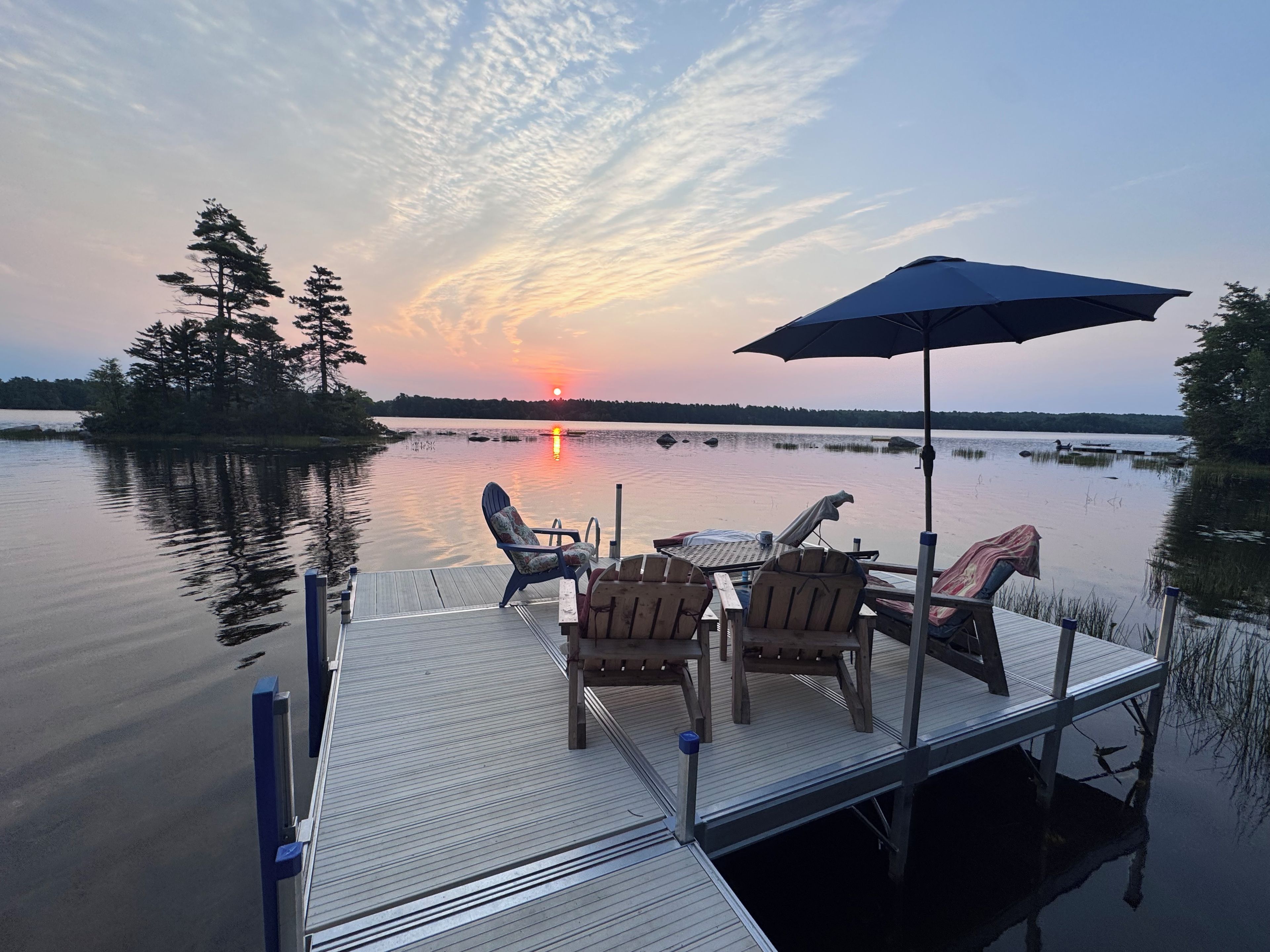 Une jetée en bois, avec des chaises et un parasol, d’où l’on peut admirer un lac paisible et un coucher de soleil.