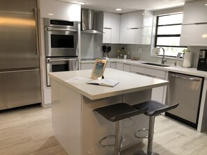 A modern kitchen with stainless steel appliances, a central island with bar stools, and a window providing natural light. - Oceanside Vacation Townhome - Sleeps 11 (Key Largo)