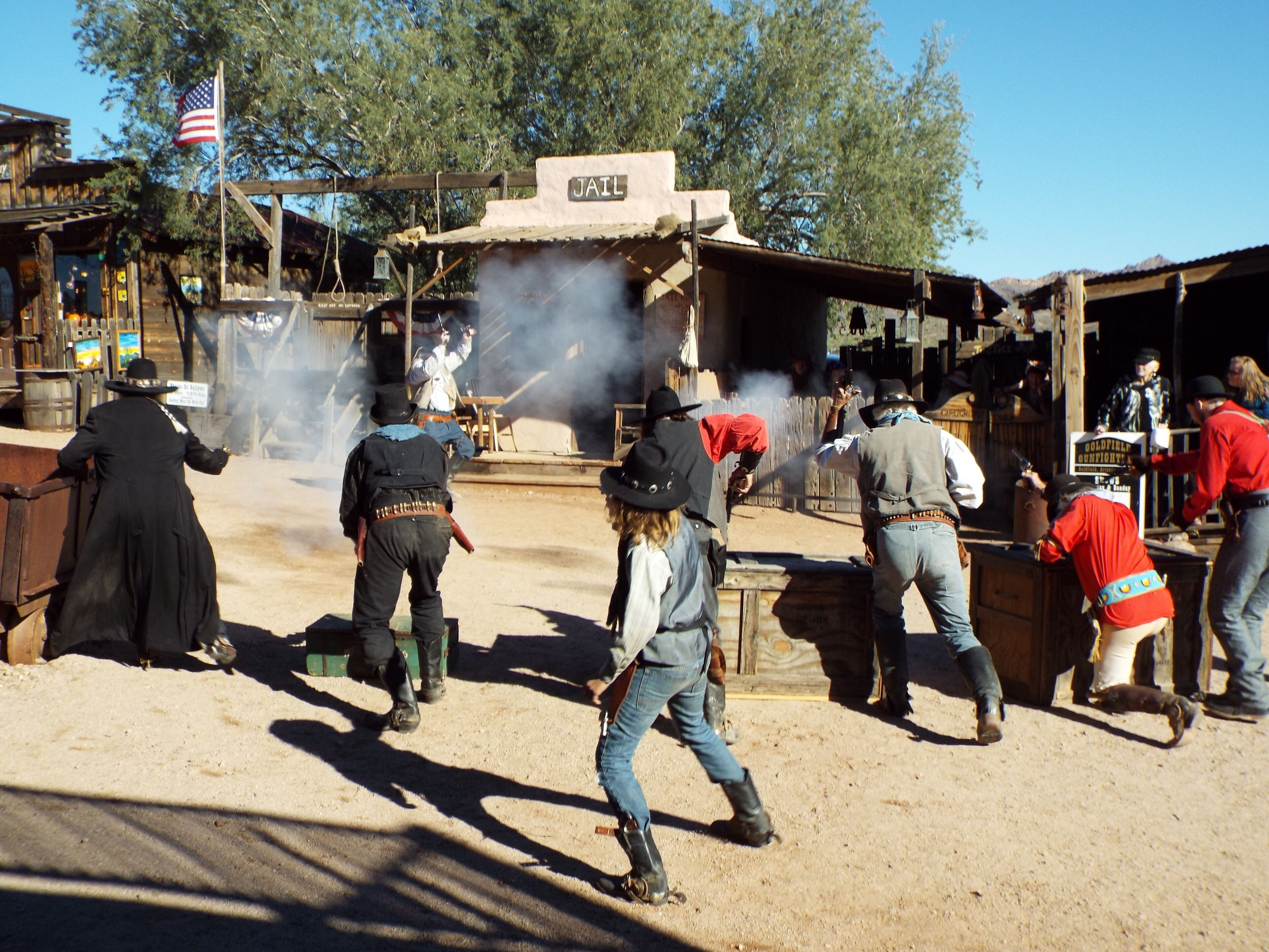 A historical reenactment scene with actors in period costumes, a wooden jail structure, and a sign indicating a goldfield gunfight event.
