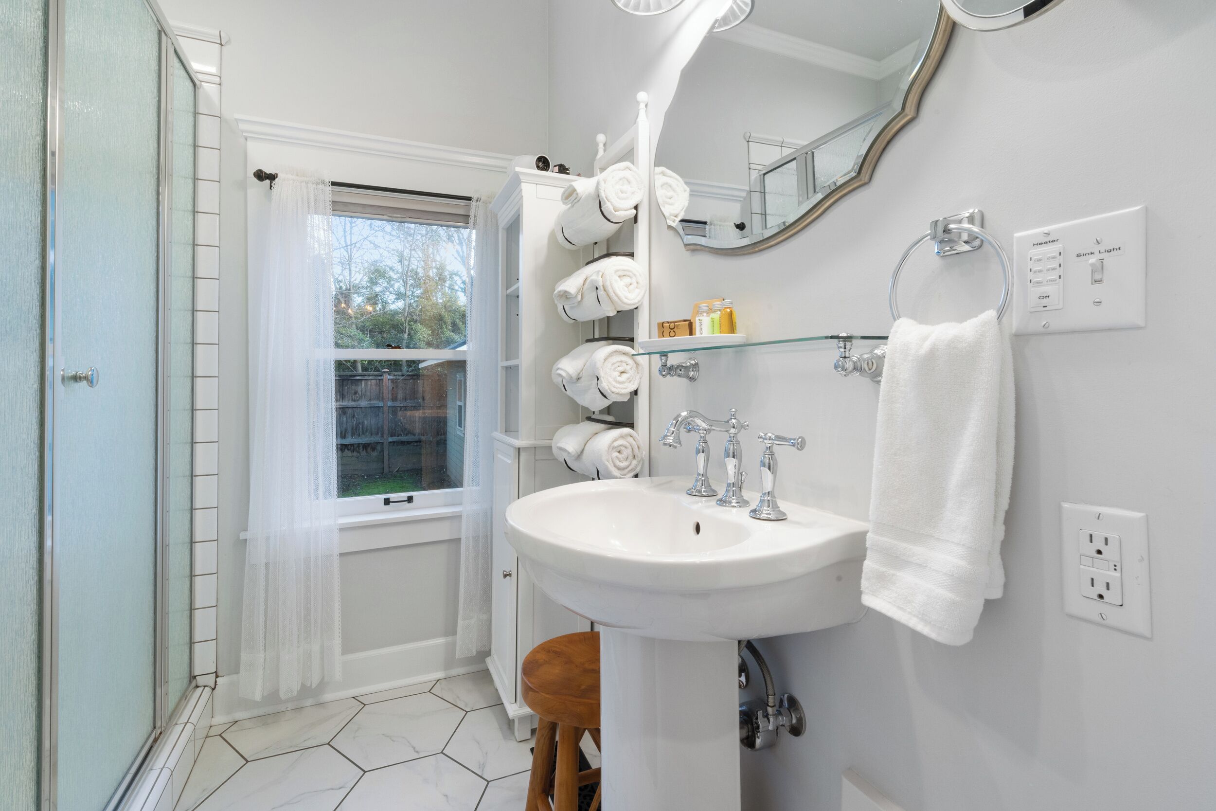 A bathroom with a white sink, a mirror, a towel rack with multiple towels, and a window with a sheer curtain.