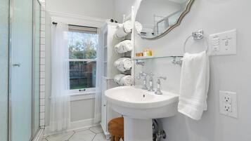 A bathroom with a white sink, a mirror, a towel rack with multiple towels, and a window with a sheer curtain.