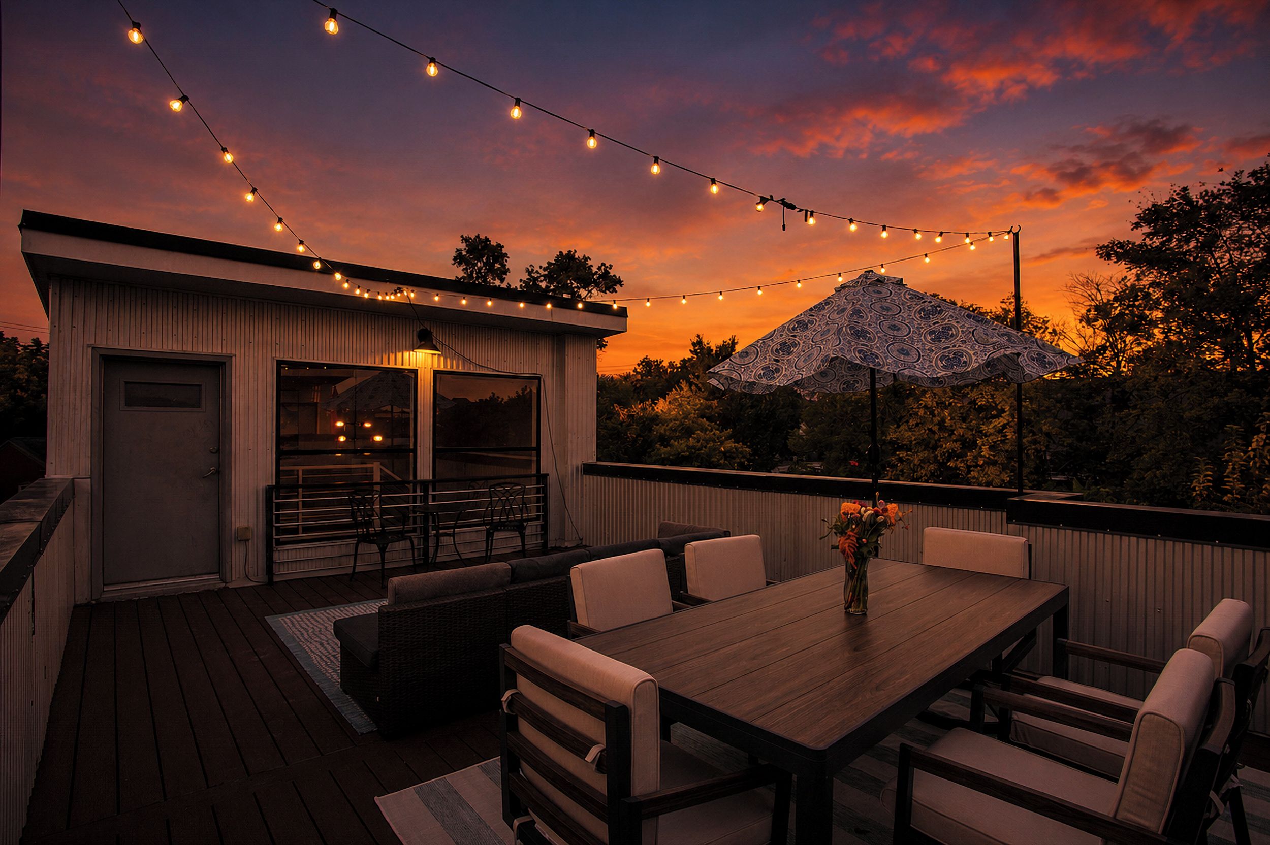 An outdoor patio with a wooden deck, a dining table with chairs, string lights, and a patterned umbrella.