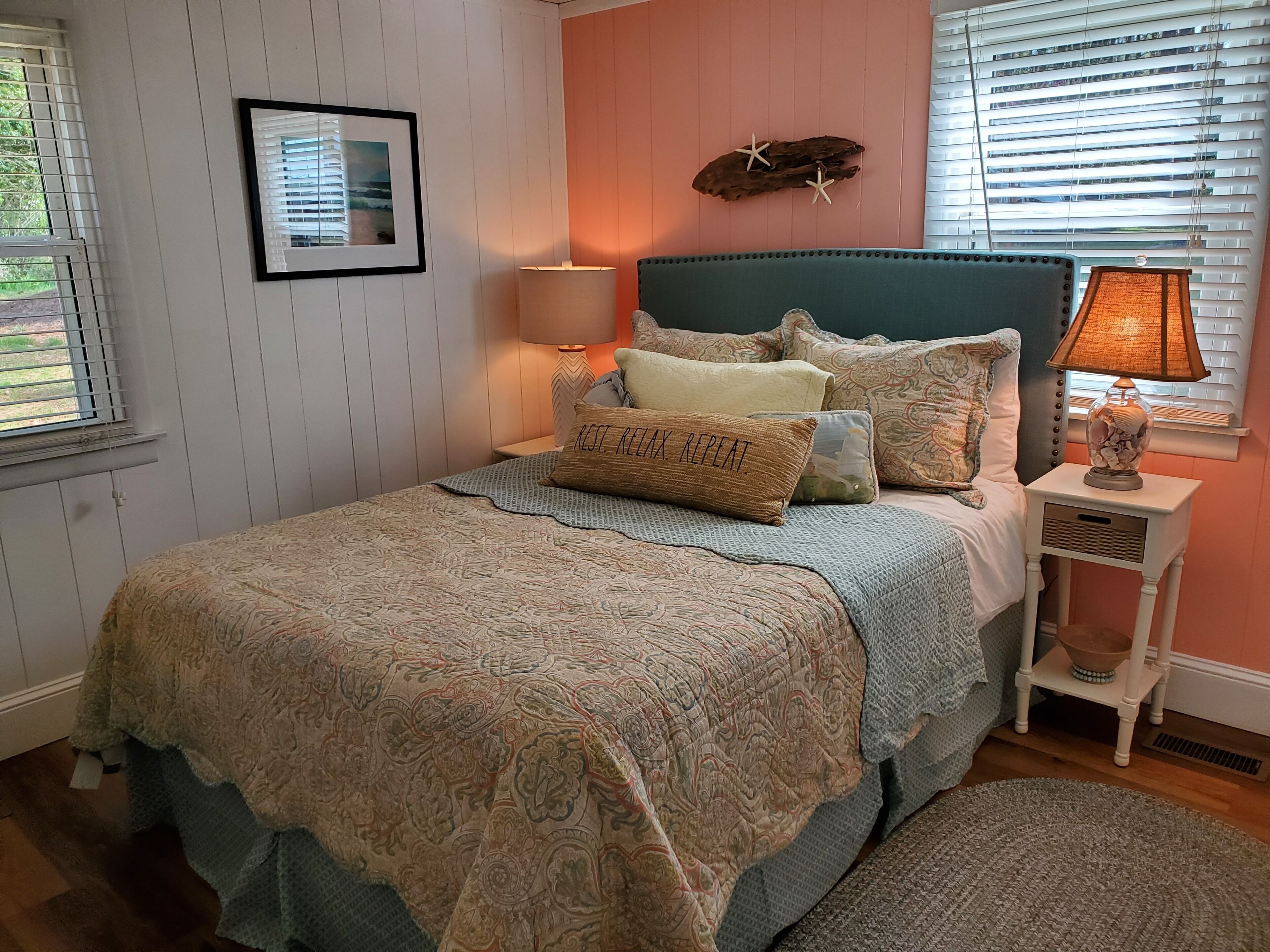 A neatly arranged bedroom with a patterned bedspread, pillows, a headboard, a bedside lamp, and a framed picture on the wall.