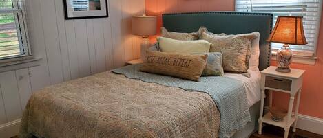 A neatly arranged bedroom with a patterned bedspread, pillows, a headboard, a bedside lamp, and a framed picture on the wall.