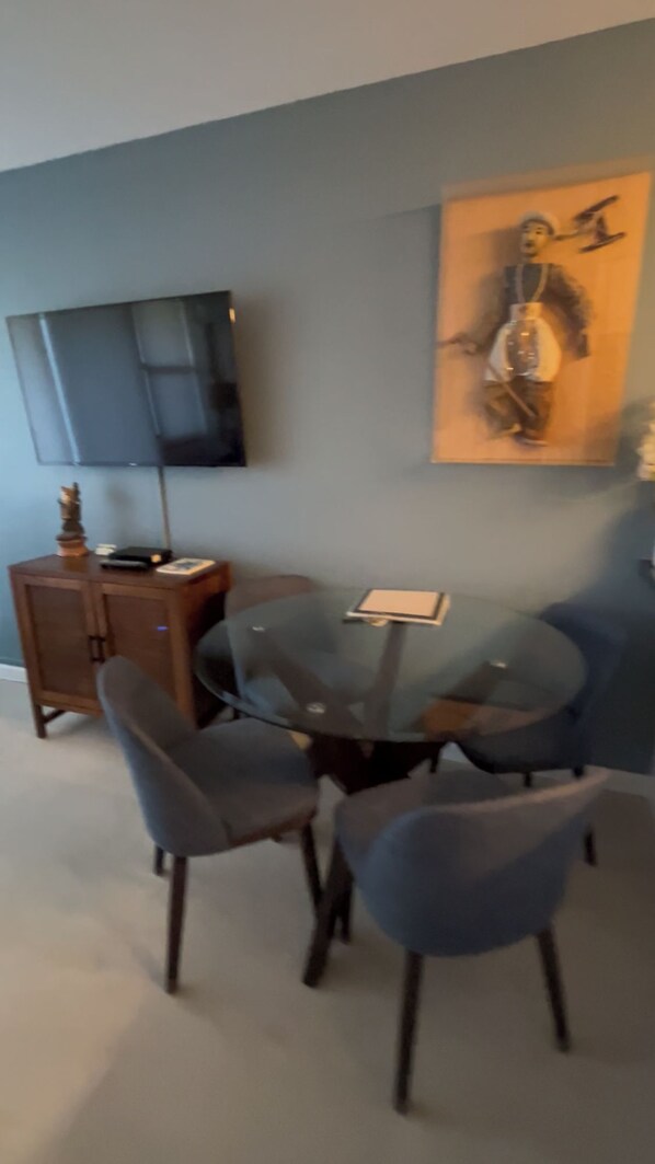 A dining area with a round glass table, four chairs, a flat-screen TV mounted on the wall, and a painting above the table. - Oooh! Oooh! Pick Us! Steps to the Beach Newly Renovated Ground Unit! (Kihei)