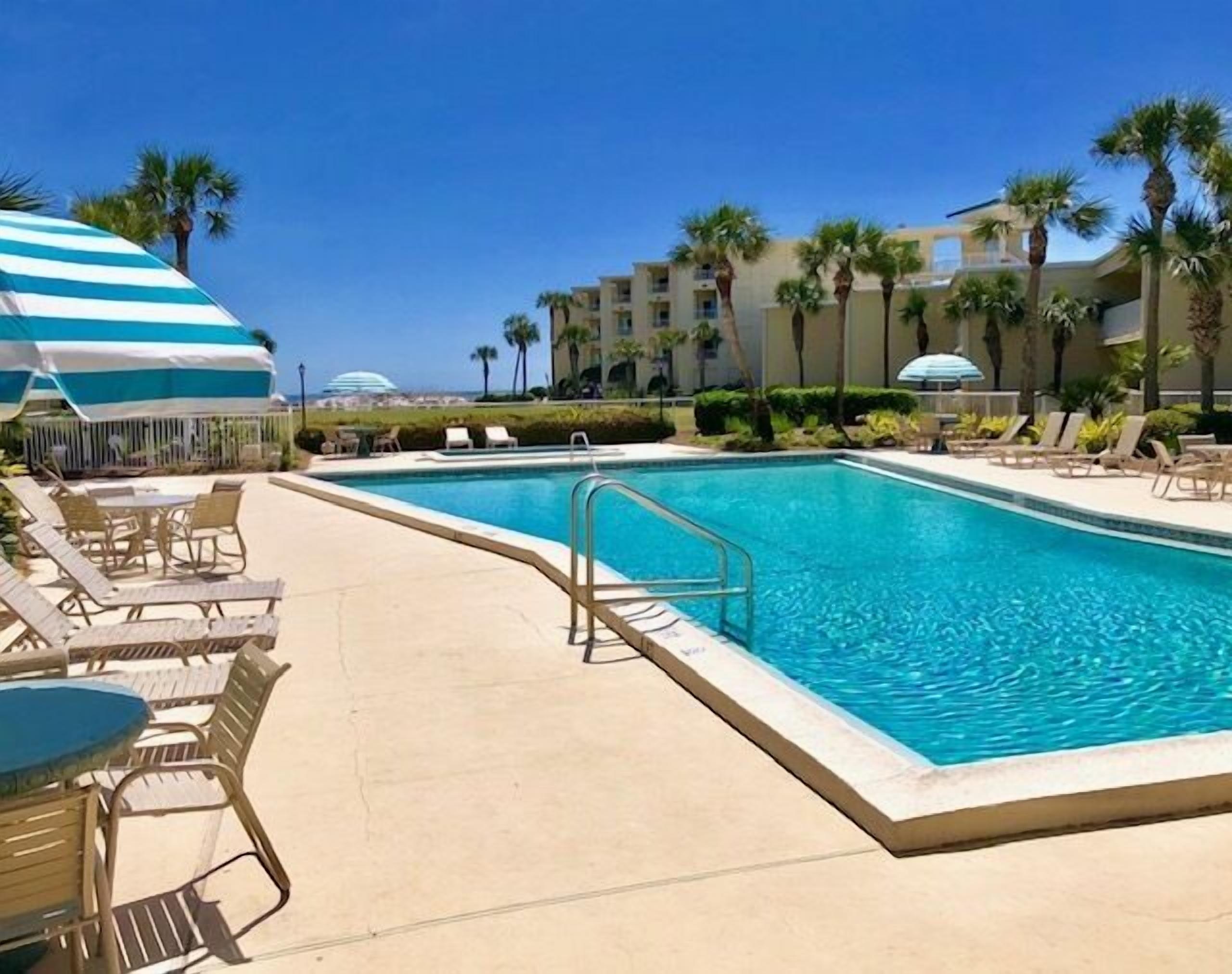 A pool area with lounge chairs and umbrellas, surrounded by palm trees and a clear sky.