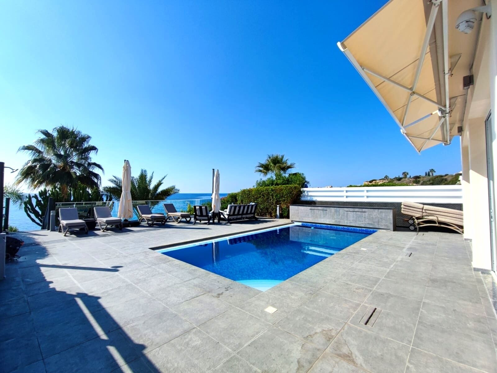 A swimming pool with lounge chairs and umbrellas, a clear view of the sea, and a white building with a balcony.