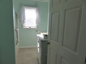 Un couloir étroit avec une porte sur la droite, une fenêtre ornée d’un rideau blanc et un radiateur sur la gauche.
