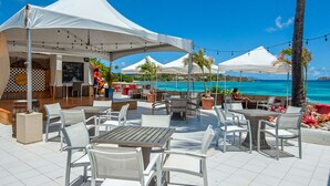 An outdoor patio with white tables and chairs, a bar area, and a view of the ocean. - Our Walk in Paradise Level at Sapphire Village, East End (St Thomas)